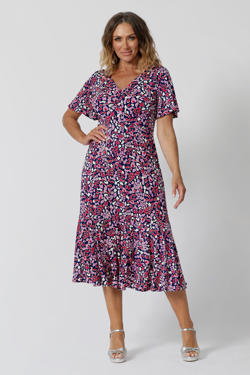 Summer dresses for over 50s women doen't get much beter than this! A size 12, over 50 woman wears an empire line midi dress with flutter sleeves and twist front bodice. A good dress for wedding guest outfits, the pink, red, white and navy blue print make it a pretty summer dress. Made in Australia by Australian and New Zealand women's clothes brand, Leina & Fleur, shop their petite to plus size dresses online.