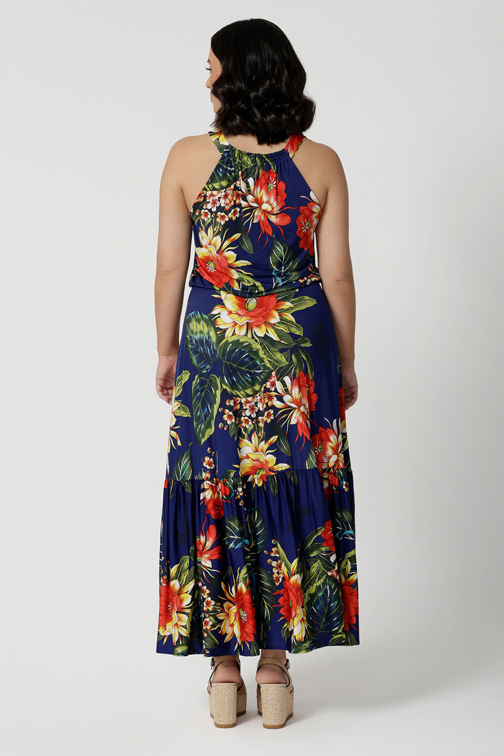 Back view of petite model wearing long floral dress with halter neck detail. Easy care dress is made of deep blue slinky jersey and has a colourful combination of orange, yellow and white flowers. Travel Ready dress comes in sizes 8-24.