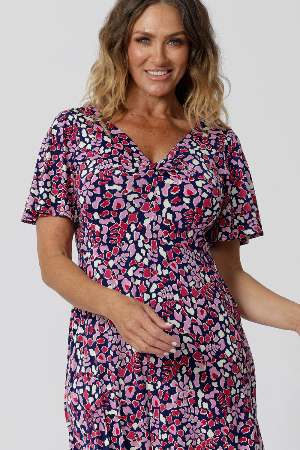 Close up of the wist front bodice on a summer dresses for over 50s women. A size 12, over 50 woman wears an empire line midi dress with flutter sleeves and twist front bodice. A good dress for cruisewear, the pink, red, white and navy blue print make it a pretty summer dress. Made in Australia by Australian and New Zealand women's clothes brand, Leina & Fleur, shop their petite to plus size dresses online.