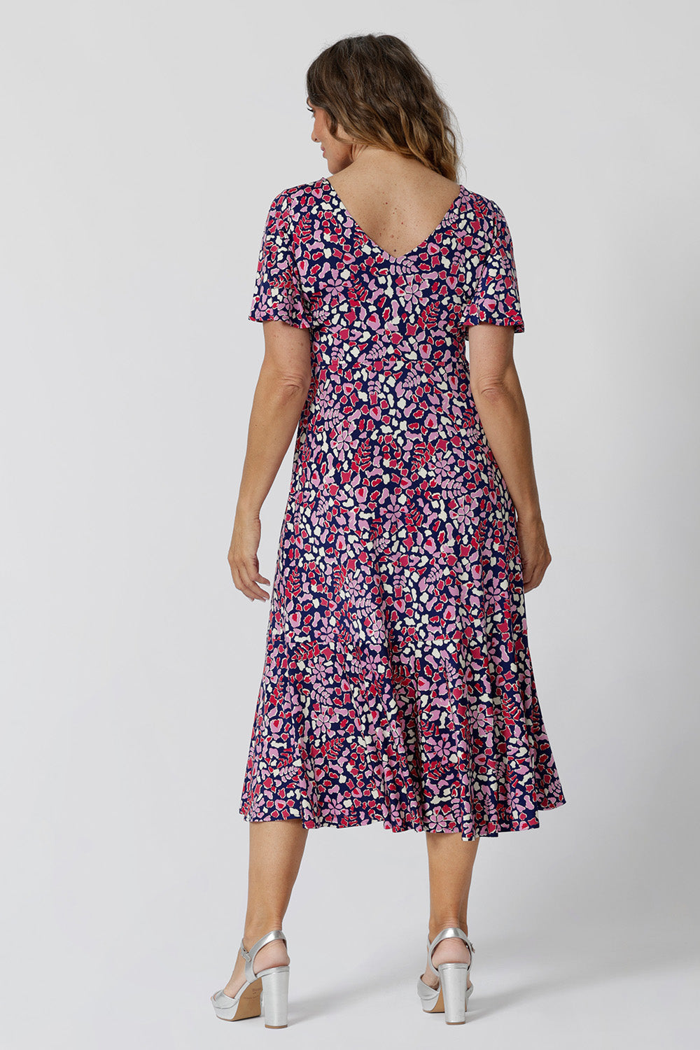 Back view of a summer dress for over 50s women. A size 12, over 50 woman wears an empire line midi dress with flutter sleeves and twist front bodice. A good dress for wedding guest outfits, the pink, red, white and navy blue print make it a pretty summer dress. Made in Australia by Australian and New Zealand women's clothes brand, Leina & Fleur, shop their petite to plus size dresses online.