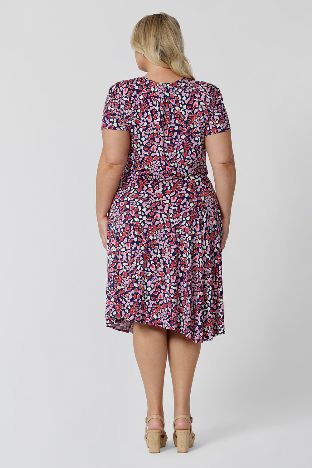 Back view of a summer dress for curves. A size 18, plus size woman wears knee length summer dress with asymmetric hemline and cowl neck. A short sleeve dress in pretty red, pink, white and navy print this is a good jersey dress for travel and cruise wear. Made in Australia by Australian and New Zealand women's clothes brand, Leina & Fleur, shop their petite to plus size dresses online.