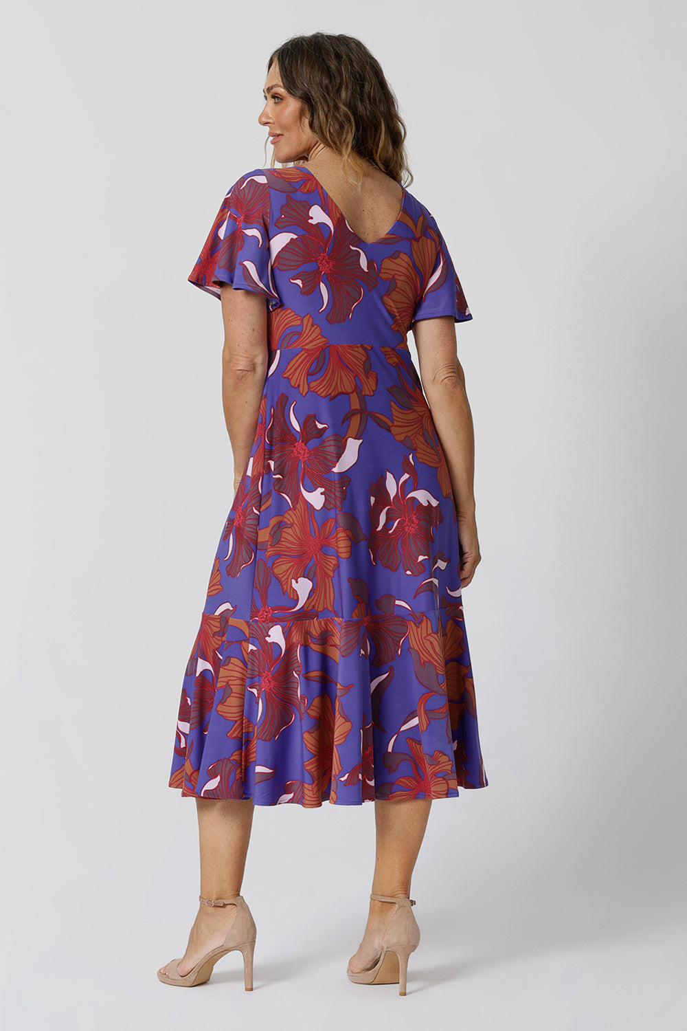 An image of size 12, over 40 woman wears an empire line midi dress with flutter sleeves and twist front bodice. She is facing backwards to show the v-neckline at the back of the dress. A good dress for wedding guest outfits, bold print makes it a pretty summer dress. Made in Australia by Australian and New Zealand women's clothes brand, Leina & Fleur, shop their petite to plus size dresses online.