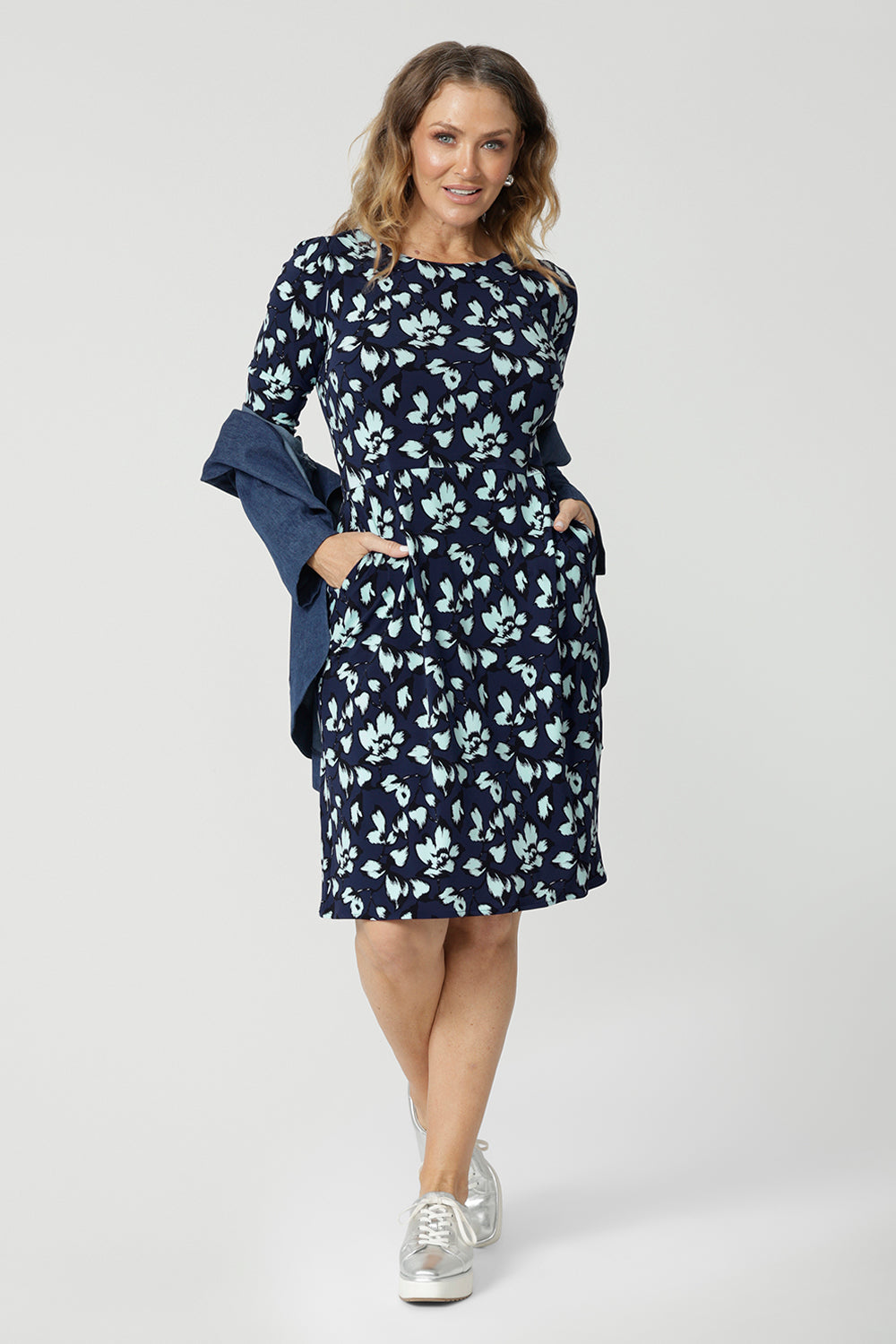 An over 40 woman wears a round neck shift dress in green and blue floral print. A good casual dress for women, this jersey dress is worn with a vintage denim jacket. Both jacket and dress are made in Australia by Australian and New Zealand women's fashion brand, Leina & Fleur. Available in sizes 8 to 24, shop this casual dress with sleeves in plus size and petite sizes.