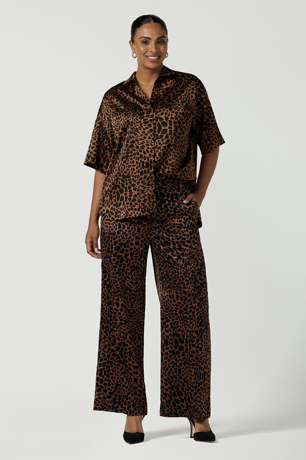 A woman wears the Sloane Pant in Bronze Animal. A satin straight leg waisted pant with pockets and a zip front. Made in high fashion Animal print fabric. Petite to plus size women size 8-24. Styled back with the Pele Shirt in Bronze animal.