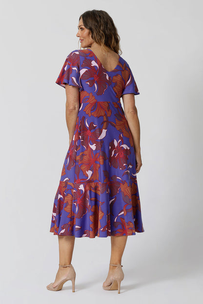 An image of size 12, over 40 woman wears an empire line midi dress with flutter sleeves and twist front bodice. She is facing backwards to show the v-neckline at the back of the dress. A good dress for wedding guest outfits, bold print makes it a pretty summer dress. Made in Australia by Australian and New Zealand women's clothes brand, Leina & Fleur, shop their petite to plus size dresses online. 