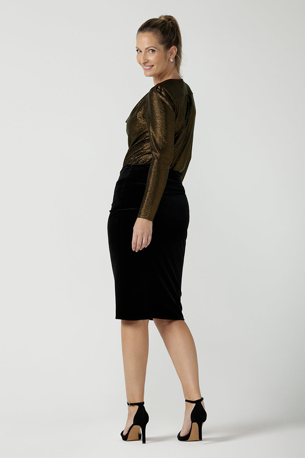 Woman wears the Brooke Skirt in Black Velour. Slim fit tube skirt for evening wear. Flattering style. Size inclusive fashion Australian made size 8 - 24. Styled back with black high heel and cowl neck gold Chrissy Top.