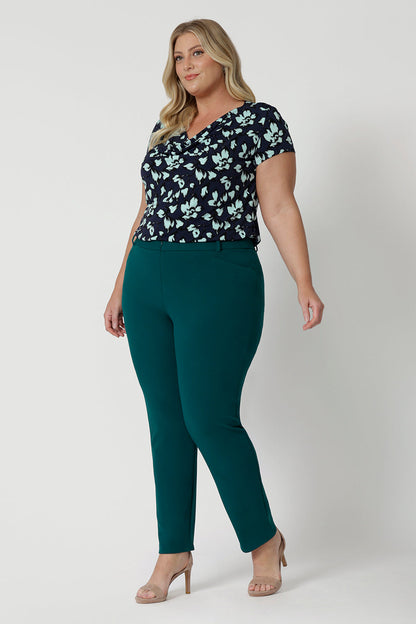 Showing a good summer top for plus size women, a curvy, size 18 woman wears a cowl neck top in aqua blue floral print. A short sleeve top , its worn with tapered leg work pants in green as  a workwear top. This comfortable top is made from stretch jersey and is an easy-care top style. Made in Australia by Australian and New Zealand women's clothes brand, Leina & Fleur.