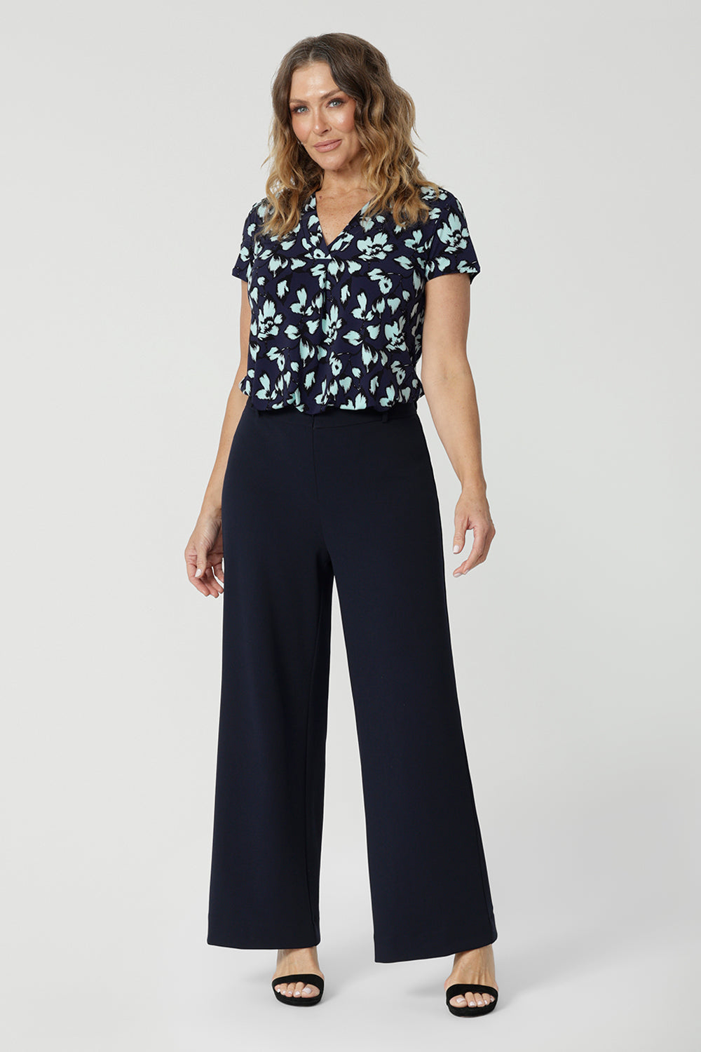 Full length shot of a good work top for women, a size 12, over 50s woman wears a short sleeve, V-neck top in aqua blue floral print. This stretch jersey top for work is worn tucked in to wide leg, Navy blue for pants for corporate outfit, Made in Australia by Australian and New Zealand women's clothes brand, Leina & Fleur, shop over 50s fashion online in sizes 8 to 24.