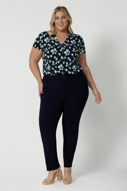 Full length shot of a good top for plus size women. A size 18, curvy woman wears short sleeve, V-neck top in aqua blue floral print. This stretch jersey top for work is worn tucked in to tapered leg, Navy blue for pants for corporate outfit, Made in Australia by Australian and New Zealand women's clothes brand, Leina & Fleur, shop curve fashion online in sizes 8 to 24.