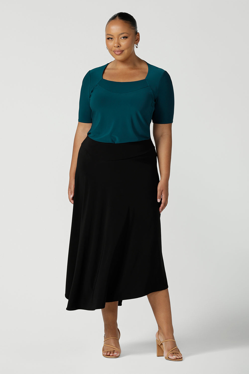 A size 18, plus size woman wears an asymmetric skirt in black jersey. Worn with a teal green short sleeve top with square neck, this comfortable jersey skirt works for work wear and eveningwear. Made in Australia by women's clothing brand, Leina & Fleur in petite, mid size and plus sizes. 