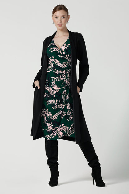 A size 10 model wears the Alpine Fern Heather dress in jersey. Made in Australia for women size 8 - 24. Fern green with leaf print all over. Styled back with the black Sorel Trenchcoat and winter boots. 