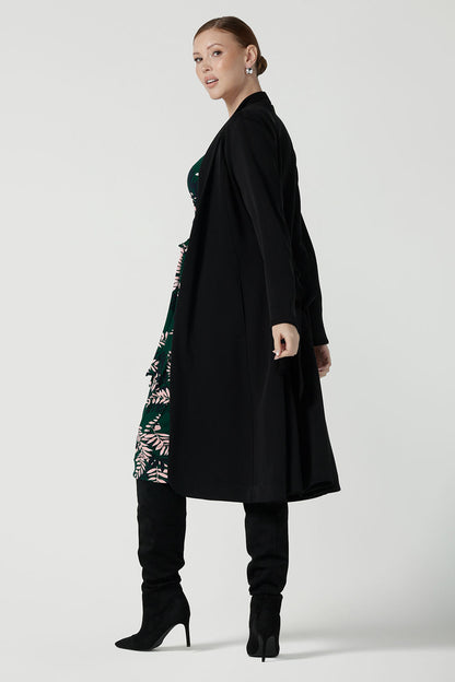 A size 10 model wears the Alpine Fern Heather dress in jersey. Made in Australia for women size 8 - 24. Fern green with leaf print all over. Styled back with the black Sorel Trenchcoat. 