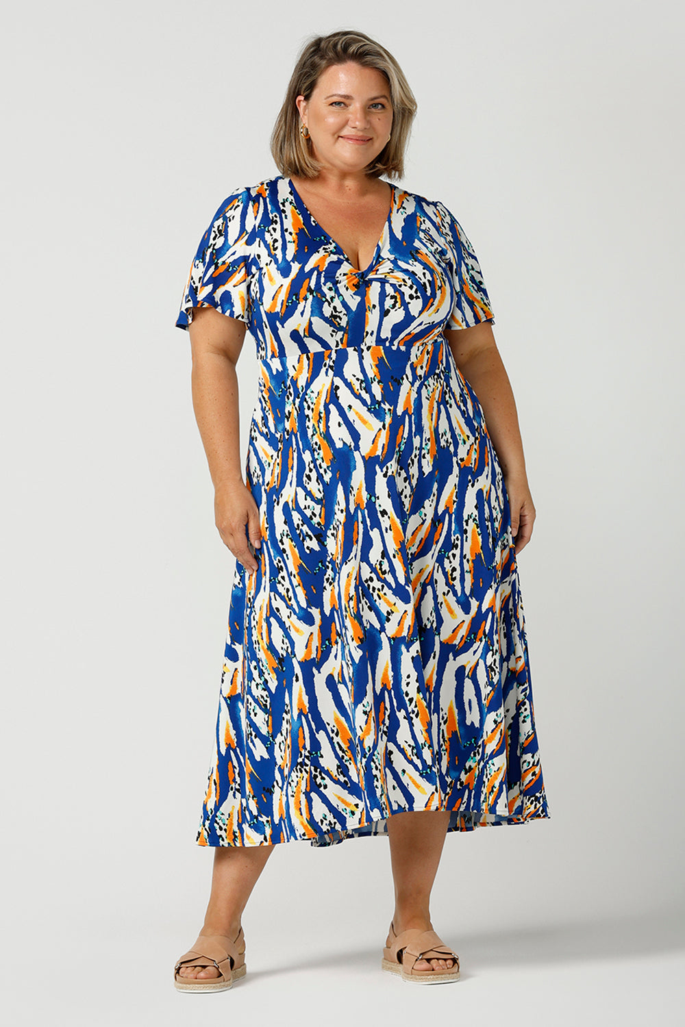 A plus size, size 18 woman wears a casual slinky jersey midi dress in an abstract print. The dress has short flutter sleeves, a double layer twist front bodice and an asymmetrical skirt - this is a great dress for weekend and travel style.