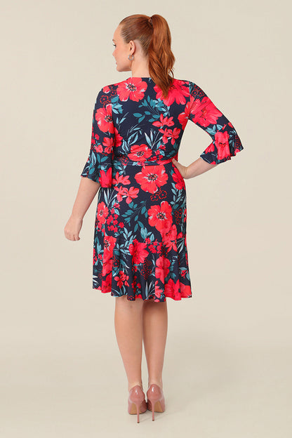 Back view of a size 12 curvy woman wearing a reversible wrap dress with 3/4 fluted sleeves. Featuring a mini-length skirt with ruffle hem and 3/4 length fluted sleeves, this floral printed jersey dress is has a v-neckline and wears well as an everyday dress. 