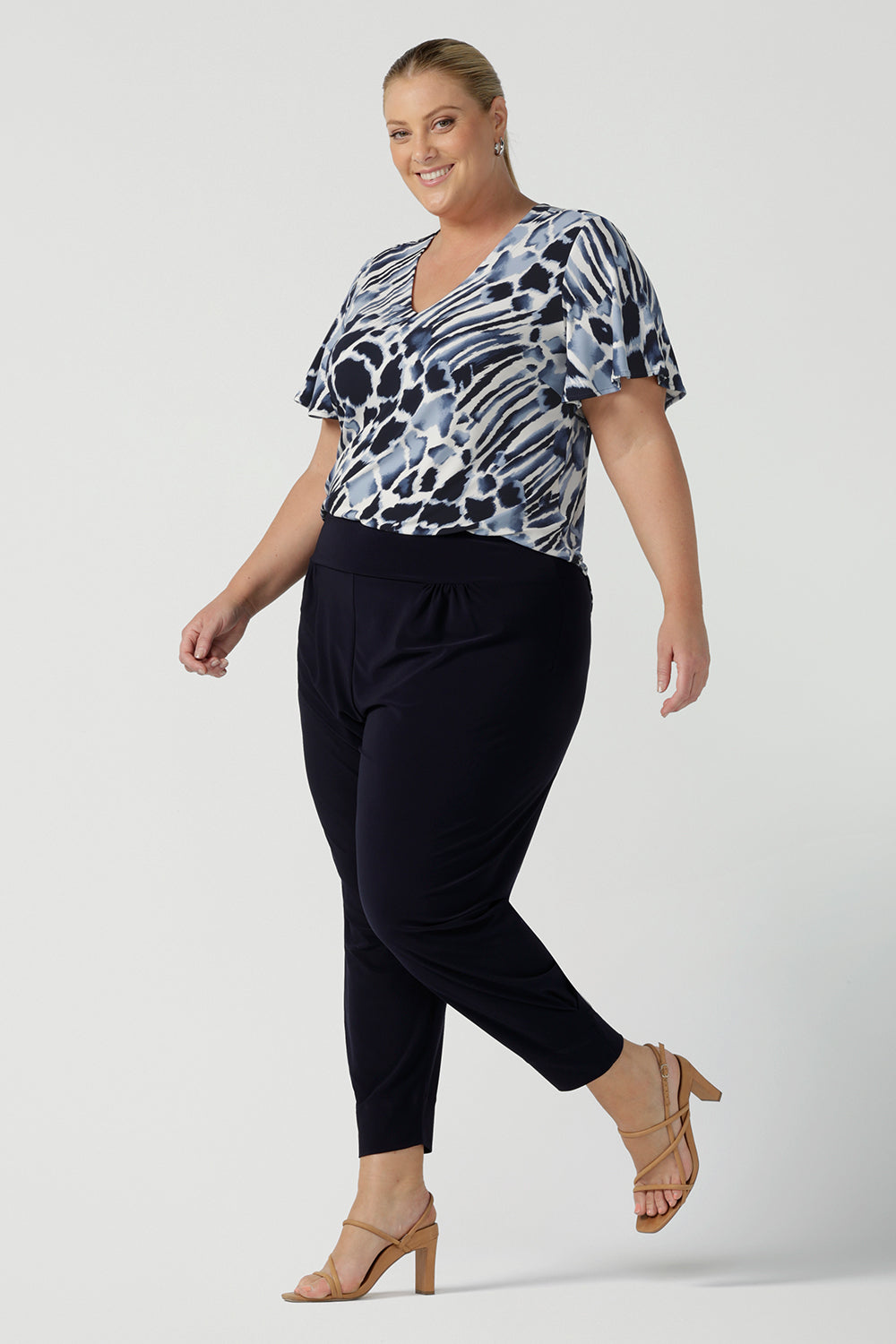 A plus size, size 18 woman wears a blue and white print jersey, V-neck top with flutter sleeves and tapered leg, dropped crotch, navy blue travel pants. A good top for summer casual wear or as a workwear top this quality women's top is made in Australia. Shop made in Australia tops in petite to plus sizes online at Australian fashion brand, Leina & Fleur.