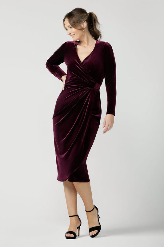 A 40 plus woman wears a long sleeve cocktail dress in wine velour. A wrap dress with midi length tulip skirt, this elegant dress is great for cocktail and eveningwear. Made in Australia by women's occasionwear brand, Leina & Fleur, shop event dresses in petite to plus sizes online and step out in style!