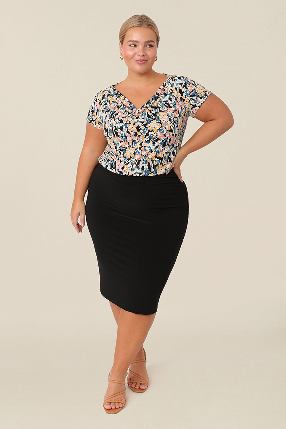 A petite height, size 14 woman wears a casual jersey top. The top has short raglan sleeves, a cowl neckline and is printed with a floral pattern paired with black tube skirt for a work wear look.