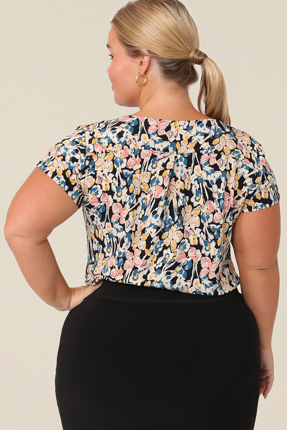 Back view of a petite height, size 14 woman wearing a casual jersey top. The top has short sleeves, a cowl neckline and is printed with a floral pattern for a casual weekend look.
