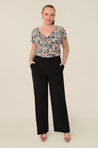 A size 12 curvy woman wears a cowl neck short sleeve jersey top in a floral print. She wears the top with wide leg trousers in black. Both are designed and made in Australia for petite to plus size women.