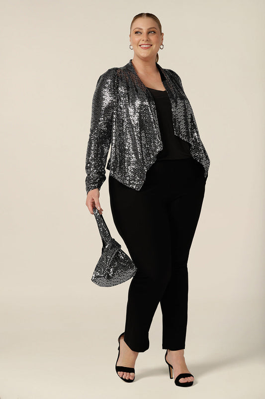 A size 18, plus size woman wears a silver sequin jacket with waterfall front for evening and event wear. Styled with a black cami top and slim leg black trousers and sequin knot evening bag, this sparkly jacket is glamorous cocktail attire at it's finest! Made in Australia by women's occasionwear brand, Leina & Fleur, shop cocktail jackets online in petite to plus sizes