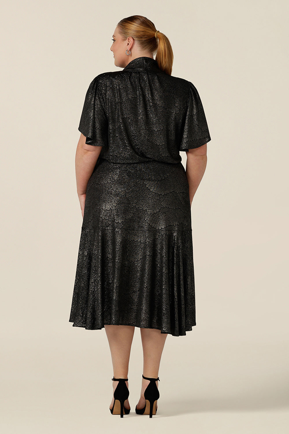 Back view of a plus size, size 18 woman wearing a good top for curvy women's evening and event wear. In shimmering bronze foil print, this V-neck top with short flutter sleeves is worn with midi length foil print skirt with ruffle hem. Styled together as a faux cocktail dress, both going out top and skirt are made in Australia by women's occasionwear brand, Leina and Fleur.