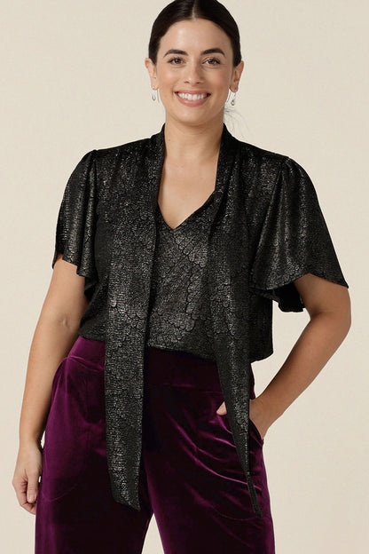 A petite height woman wears a good top for women's evening and event wear. In shimmering bronze foil print, this V-neck top with short flutter sleeves is made in Australia by women's occasionwear brand, Leina and Fleur.