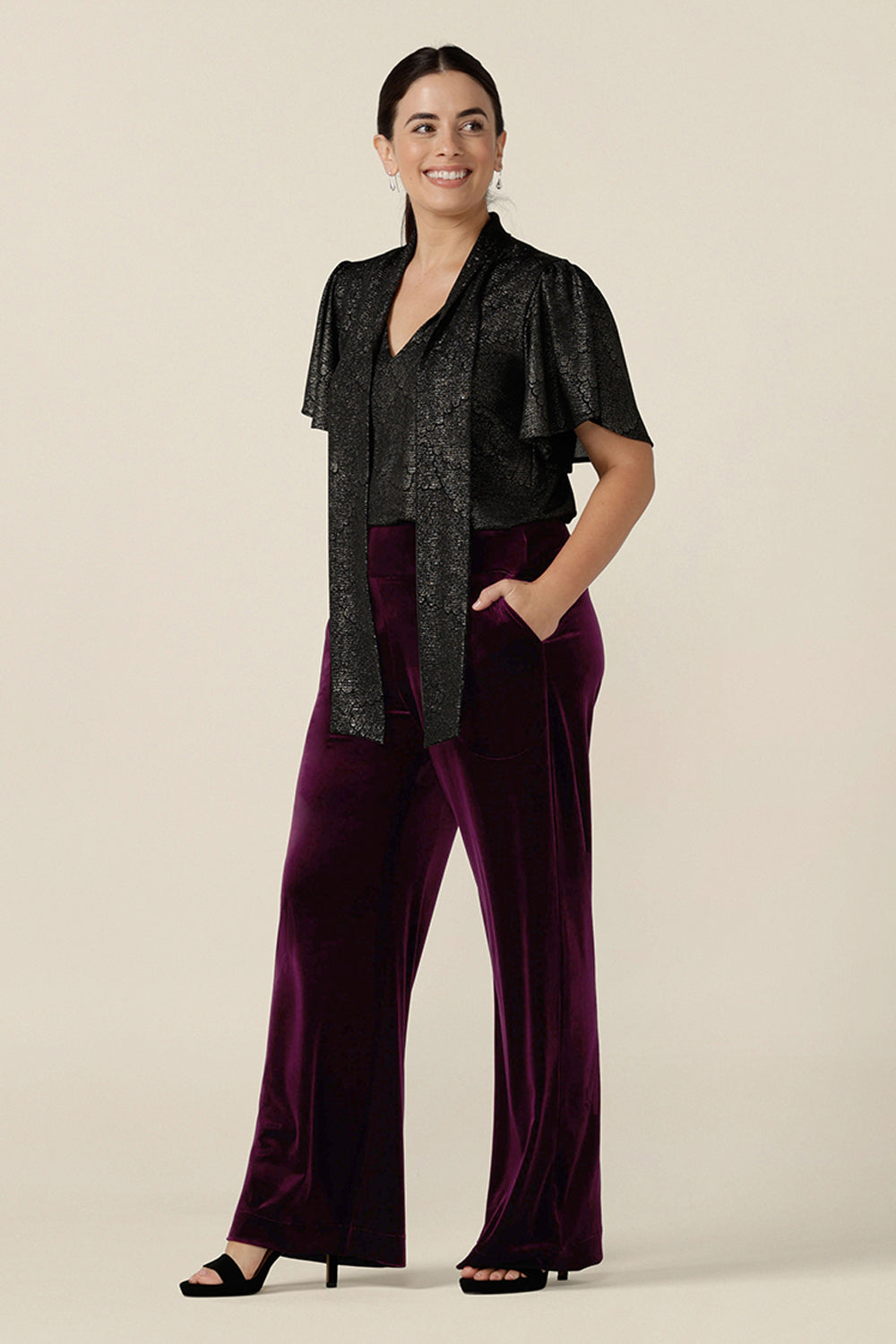 A petite height woman wears a good top for women's evening and event wear. In shimmering bronze foil print, this V-neck top with short flutter sleeves is worn with wide leg velvet evening trousers. Both evening top and cocktail pants are made in Australia by women's occasionwear brand, Leina and Fleur.