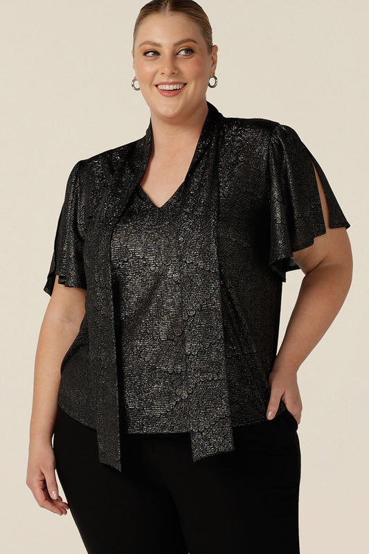 A plus size, size 18 woman wears a good top for curvy women's evening and event wear. In shimmering bronze foil print, this V-neck top with short flutter sleeves is made in Australia by women's fashion brand, Leina and Fleur.