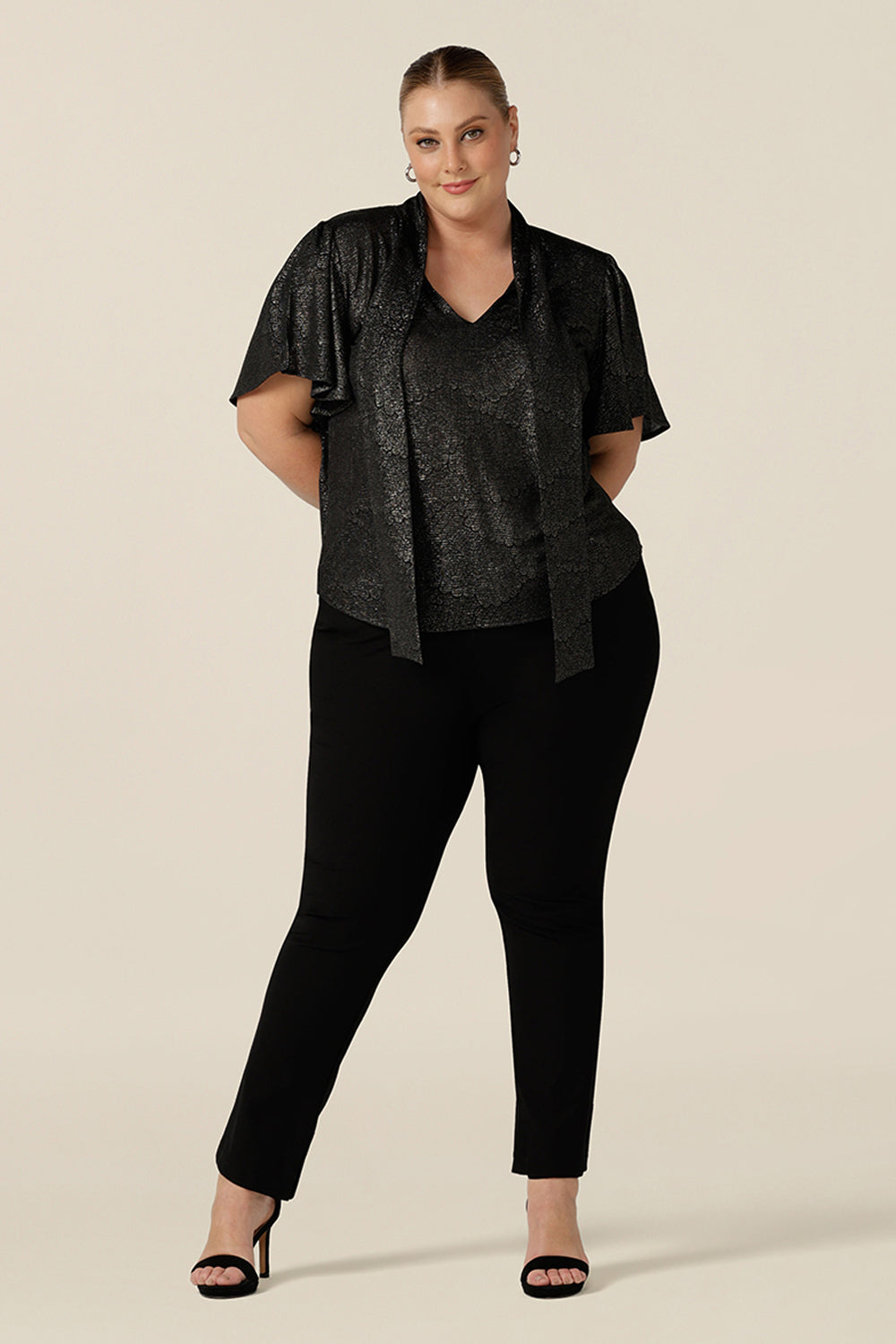 A plus size, size 18 woman wears a good top for curvy women's evening and event wear. In shimmering bronze foil print, this V-neck top with short flutter sleeves is worn with slim leg black trousers for an elegant cocktail wear look.  Made in Australia by women's occasionwear brand, Leina and Fleur.