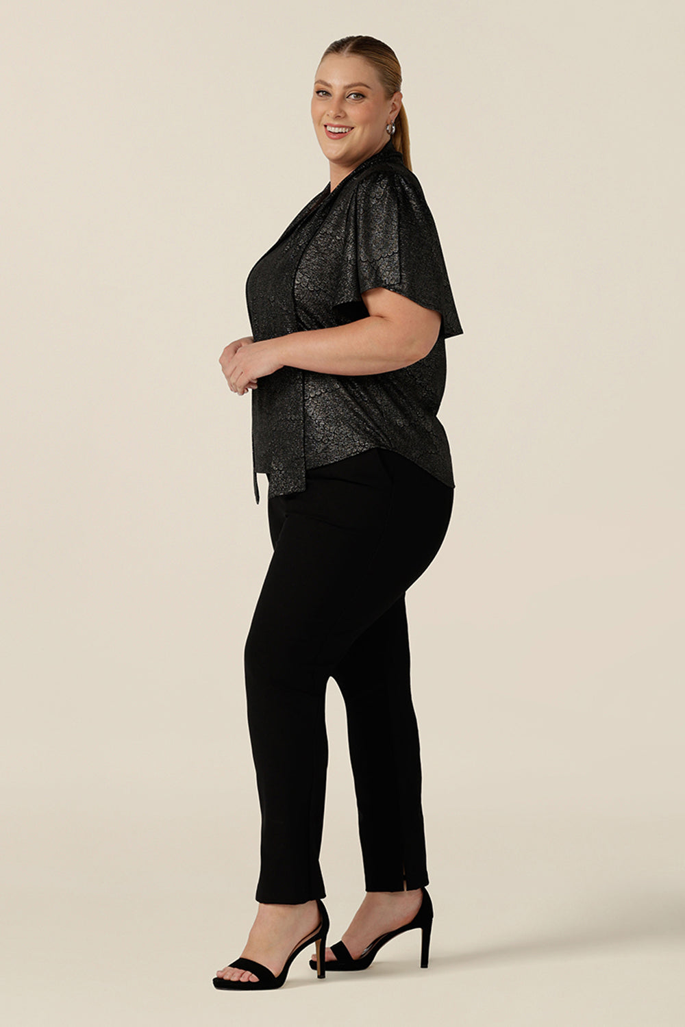 Side view of a plus size, size 18 woman wearing a good top for curvy women's evening and event wear. In shimmering bronze foil print, this V-neck top with short flutter sleeves is worn with slim leg black trousers for an elegant cocktail wear look. Made in Australia by women's occasionwear brand, Leina and Fleur.