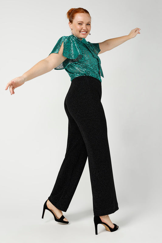 A woman wears sparkly black pants for cocktail and evening wear. In size 12, these wide leg cocktail wear pants are great plus size summer party pants for dazzling curves! Worn with a jade metallic flutter sleeve top, shop wide leg cocktail pants in petite to plus sizes at Leina & Fleur.