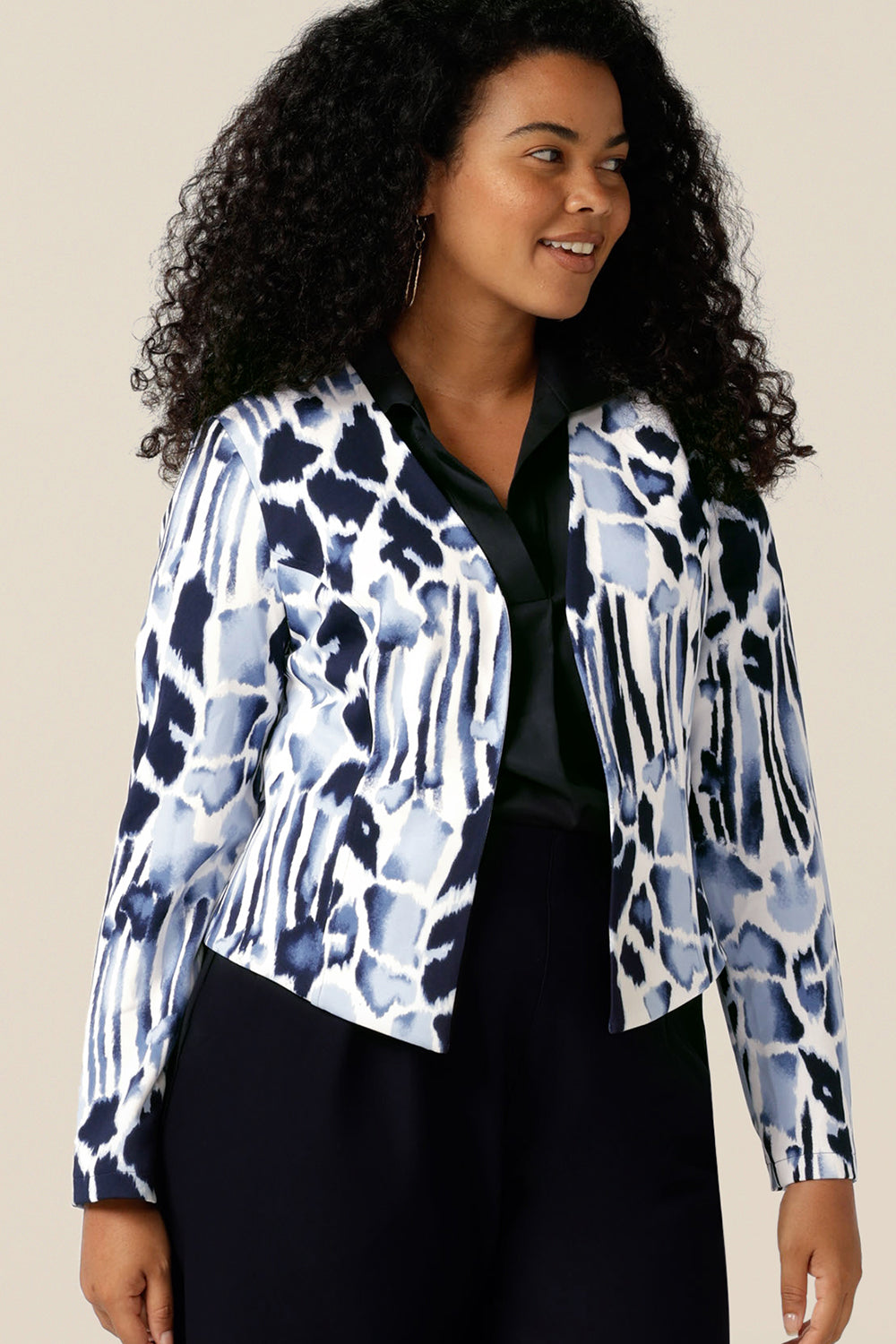A size 12, curvy woman wears a collarless open front jacket for work and corporate wear. In navy and white jersey, this is a tailored jacket with stretch for all-day wear as a comfortable office jacket. Styled with a navy blue shirt and navy pants, this women's corporate jacket completes the work wear outfit.